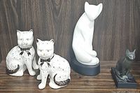 Four cat figurines arranged on wooden surface against dark wood background: two vintage Staffordshire style cats, one white abstract Li-Ching cast resin cat, one small black cat figurine.