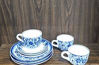 Stack of 3 blue and white ceramic dinner plates, 3 saucers and 3 mugs with floral and geometric patterns against a wooden background.