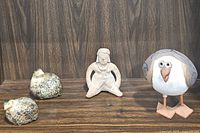 Three figurines displayed against a wood background: two small speckled quail birds, a stone pottery figurine of a person, and a ceramic seagull figurine.