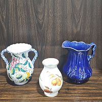 Overall view of three items included in the lot: Italian hand painted ceramic utensil holder vase, Royal Worcester Evesham porcelain vase, and cobalt blue water pitcher on wooden surface with dark wood panel background.