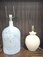 Two lamps on a wooden shelf; left is a tall modern glass lamp with a cloudy marbled effect in white and light green, brass socket, and lamp harp; right is a shorter vintage cream scalloped lamp with brass socket.