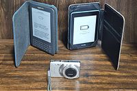 Photo showing two Amazon Kindle e-readers, one in a gray cover and one in a black folio with card slots, plus the silver OM SYSTEM OLYMPUS Stylus 7010 digital camera in front.