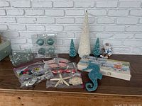 Full view of lot including stoneware plates in box, ceramic Santa figure, seashell Christmas trees, blue seahorse, multiple fish and starfish ornaments, beach sand ornament, and glass fish pieces arranged on wooden surface.