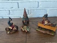 Four distinct figurines grouped on a wooden surface against a white brick wall: a gnome, hobo, upside down person, and small sitting girl figurine.