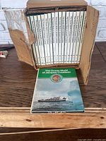 Full set of 14 hardcover books titled The Ocean World of Jacques Cousteau by Danbury Press, displayed in a cardboard box with one book standing in front showing a ship and iceberg on the cover.