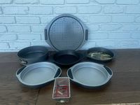 Overall view of bakeware lot showing various pans including cake pans, mini loaf pans, oval pans and pizza pan with box of skewers in front