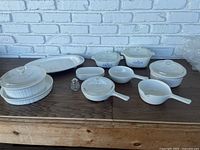 Full lot view on wooden table with white brick background showing various Corning Ware and related kitchenware items including covered cooking containers, serving platter, side dishes, and salt shaker