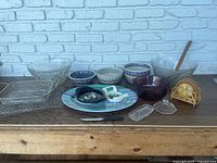 Various Polish ceramic bowls with traditional blue and white patterns, glass bowls, plastic platter, metal strainers, a glass butter dish with lid, wooden napkin holder, and kitchen tools arranged on a wooden surface against a white brick wall.
