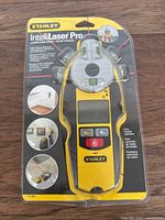 Front view of the Stanley Intellilaser Pro device in original sealed packaging showing yellow and black handheld laser level and stud finder with control buttons and bubble level indicators.