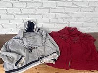 Gray zip-up hoodie and red button-down shirt laid flat on table with white brick backdrop
