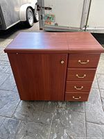Closed view of the wooden sewing machine table showing four side drawers and one cabinet door with brass handles.