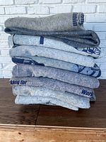 Stack of 10 used shipping blankets in various shades of grey with faded blue lettering, folded and stacked on a wooden surface by a white brick wall.