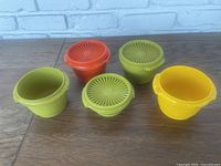 Set of five colorful plastic Tupperware storage containers with some vented lids displayed on a wooden surface against a brick background.
