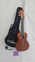 Cordoba ukulele with soft padded case, clip-on tuner, and two picks arranged in front of the case.