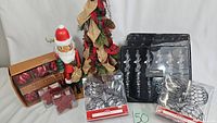Photo of assorted Christmas decorations including a small tree with plaid and burlap ribbons and pinecones, a Santa figurine, red and silver ornaments, Tahari brand packaging.