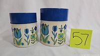 Front and slightly angled view of two vintage metal canisters with floral design and blue lids. Yellow lot number card with '57' visible.