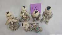 Five ceramic Asian baby figurines arranged on white surface with purple label in background