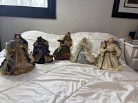 Full view of all five handcrafted paper nativity figures arranged on a white bedspread in front of a window.