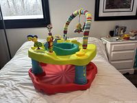 Yellow and red infant exer saucer activity center with blue legs, green padded seat, an arched toy bar with hanging soft toys, and various interactive toys fixed around the seat area.