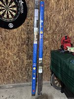 Two boxed Coolaroo outdoor roller shades, each 6x6 ft, leaning against a wooden wall next to a dartboard.