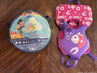Purple and pink Stearns Infant Puddle Jumper life jacket with fish graphic on wooden surface alongside the float packaging.
