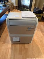 Front view of the portable air conditioner unit sitting on a wooden floor with the user manual placed on top of it.