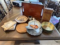 Assorted kitchen items arranged on wooden table showing serving tray, plates, wooden board, measuring spoons, knife block, and round brass container.