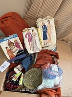 Box displaying a variety of assorted items including vintage patterns, yarn skeins, textile fabric, ribbons, and a metal dish.