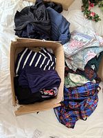 Clothing items packed loosely with some folded in a cardboard box and other garments laid out on white surface. Includes striped shirt, purple corduroy pants, floral print, and plaid pattern garments.