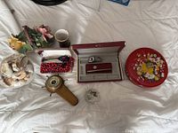 Photo showing assorted vintage items including faux porcelain flowers, seashell bowl, brown cup, red jewelry boxes with watch and clips, wooden barometer with compass, bead necklaces and floral decorated tin