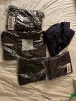 Five pieces of new clothing laid out on a bed, many in original plastic packaging, showing dark colors such as black and navy, assorted styles and sizes.