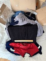 Top view of open box containing assorted clothing including a red jacket, black and gray clothing items, plus clear plastic and wooden hangers on a white fabric surface.