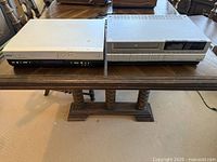 Photo showing a silver DVD player and a gray VHS player side by side on a wooden table