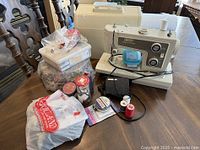 Vintage Kenmore sewing machine sitting on a wooden table with hard carrying case in background. Various sewing supplies including spools of thread, buttons in plastic bags, and storage boxes arranged nearby.