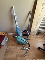 Photo showing two vintage vacuums including an upright and a canister model with hoses and attachments placed on wood floor by wall.