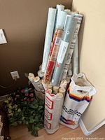Photo showing multiple wallpaper rolls stacked upright near a corner with some artificial greenery on the floor.
