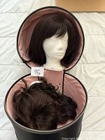 Long brown wig on white styrofoam mannequin head inside open black round two-tiered storage case with pink lining, with two additional wigs inside the case.