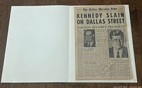 Open newspaper showing the front page with headline 'Kennedy Slain on Dallas Street'
