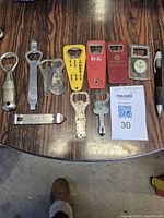 Full view of 11 assorted bottle openers laid out on wooden surface showing various styles and materials.