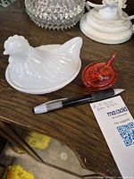 Two vintage laying hen dishes shown together with pen for scale. One is white milk glass, textured, no chips or cracks. The other is small red glass, also without chips or cracks.