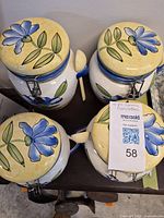Four ceramic canisters with painted blue flowers and green leaves, each with a yellow lid featuring a flower design and metal clasp. Includes matching ceramic spoons.