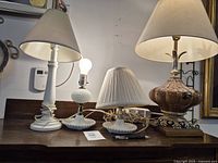 Four lamps displayed on a table; two white ceramic lamps with beige shades and two vintage milk glass lamps with pleated shades visible.