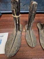 Four cast iron shoe forms positioned standing upright on a wooden surface, showing front and top views of the shoe shapes and handles. One has a rope tied on the handle.