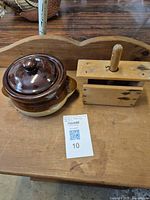 Both items shown side by side on wooden surface: ceramic brown pot with lid and wooden butter press.