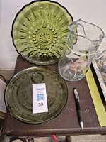 Photo showing two green glass serving plates, one standing upright with floral pattern design, and the other laying flat with auction tag. Clear glass juice jug with floral design is positioned next to plates.
