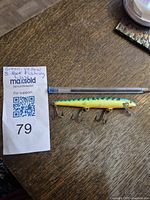 Photo of fishing lure placed on a wooden surface next to a pen for size reference and lot tag number 79 with handwritten note describing the lure as 'Green-Yellow 3-Hook Fishing Lure'