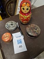 Four items displayed on a flat surface including a wooden Russian nesting doll in red and floral paint, a round pewter box with an eagle design, a small round wooden box with floral decoration, and a carved wooden box with geometric pattern.