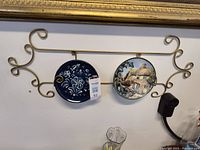 Gold colored metal wall plate rack holding two ceramic plates with distinct designs hung on a wall.