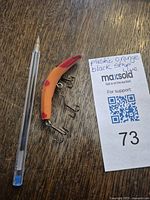 Photo showing the orange and black plastic fishing lure next to a pen and auction tag for size reference.