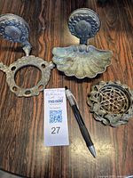 Three brass vintage bathroom wall fixtures displayed on a wooden surface with a pen and auction lot card for scale. Visible items include a mount plate, a shell-shaped soap dish holder, and ornately detailed drain cover.